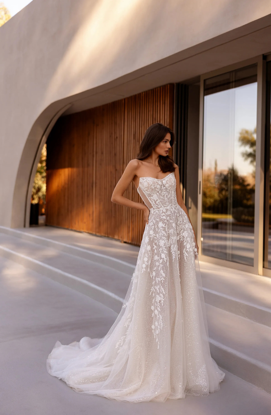 A model wearing a strapless, intricate wedding gown with detailed embroidery, standing in front of a modern architectural backdrop.