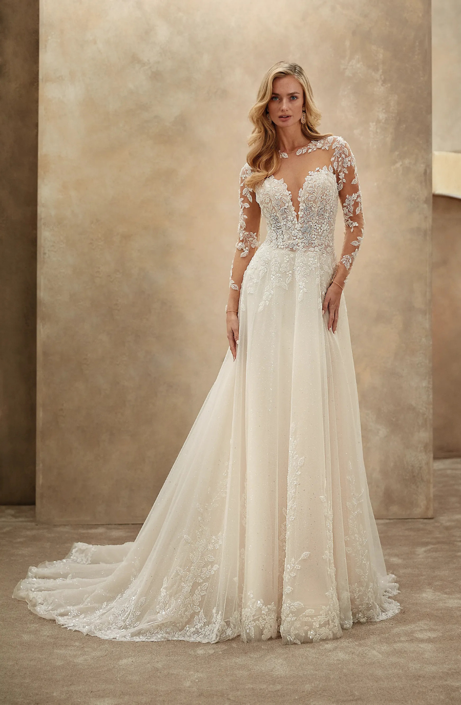 A model wearing a beautiful long-sleeved wedding dress with intricate lace details and a plunging neckline. The dress is flowy and has a train, set against a neutral background.