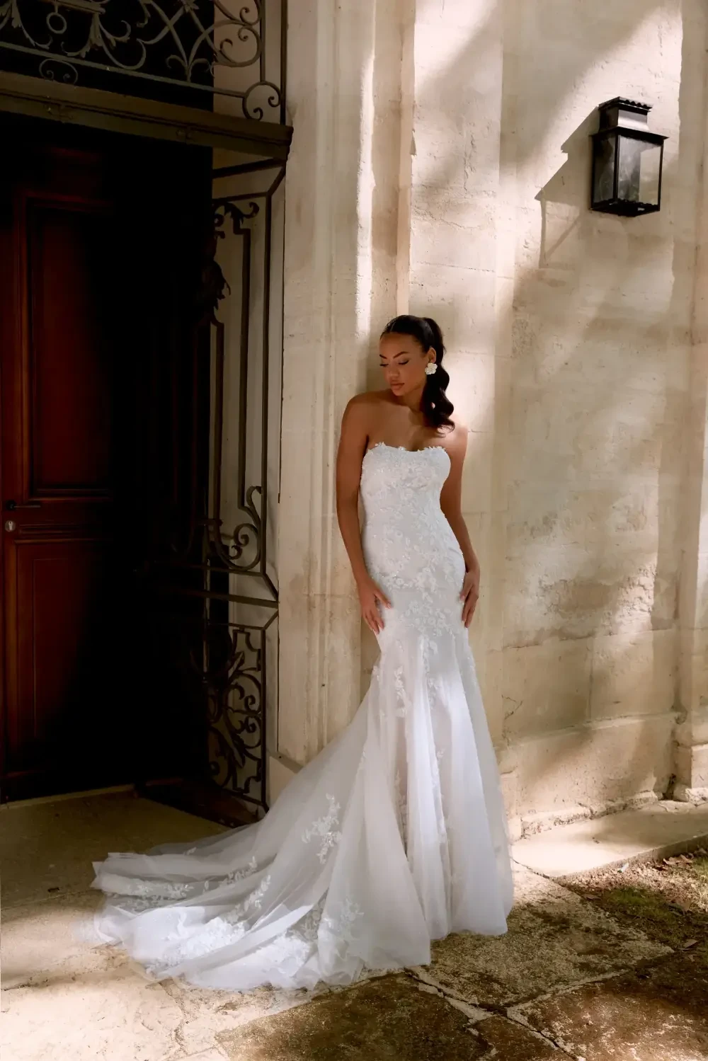 A woman in a strapless wedding gown stands against a stone wall, with a wooden door and a lantern in the background.