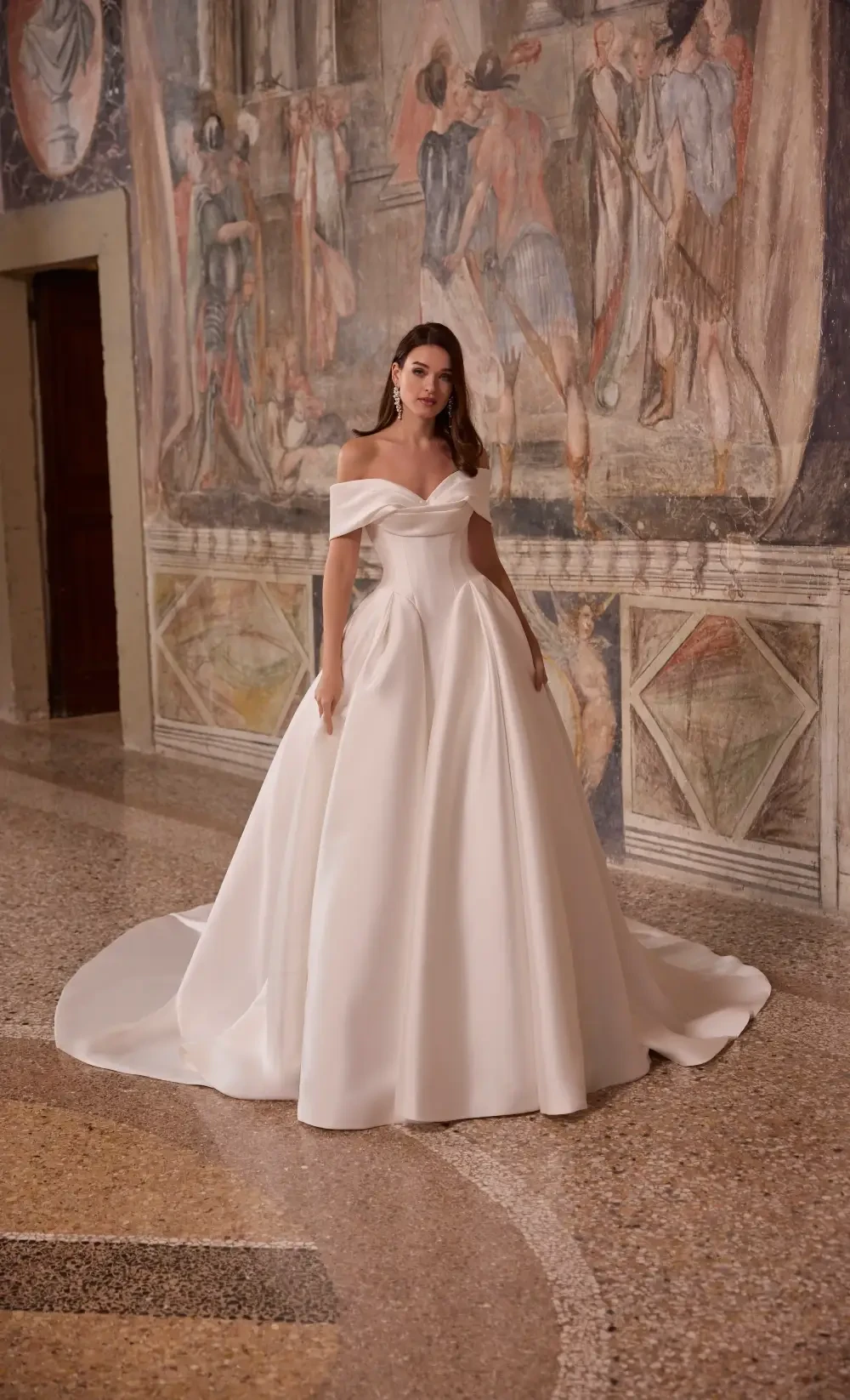 A woman in a luxurious off-the-shoulder white wedding gown stands elegantly, with a grand mural in the background.