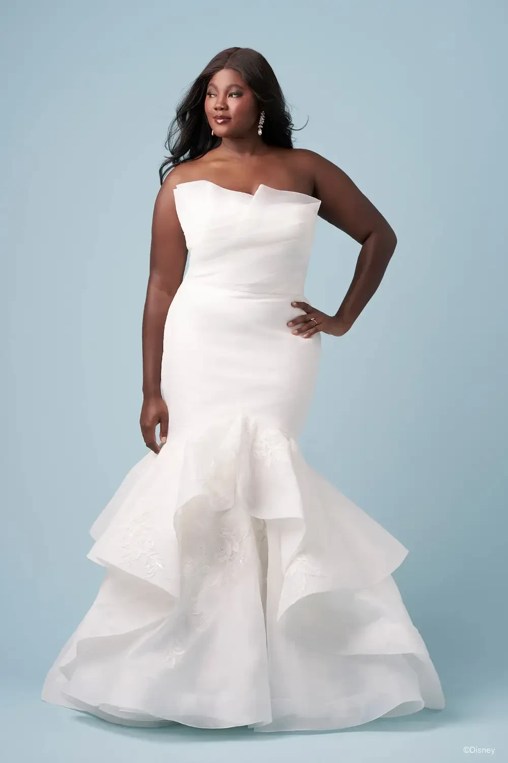 A person wearing a white strapless gown with a mermaid silhouette and layered ruffles, posing against a light blue background.